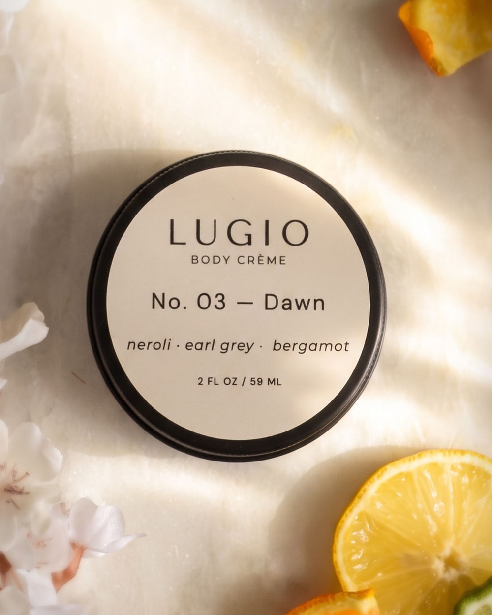 Lugio No. 03 Dawn body crème tin on white marble in warm morning light, with sliced lemon, orange and bergamot and soft white blossoms.